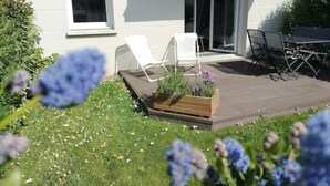 A modern patio with a wooden deck, white lounge chairs, and a table with chairs. - Logement 1 à 6 P- 150m Plage- Jardin et Parking (Courseulles-sur-Mer)