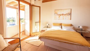 Une chambre à coucher comprenant un lit, une table de chevet, une chaise, un tableau et une porte-fenêtre coulissante donnant sur un balcon.