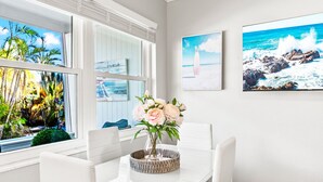 A dining area with a white table and chairs, a vase with flowers, and a view of the ocean through large windows.