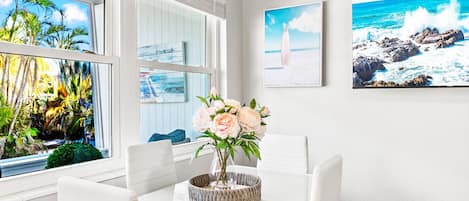 Un coin repas avec une table et des chaises blanches, un vase rempli de fleurs et une vue sur l’océan grâce à de grandes fenêtres.