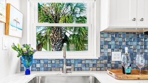 A kitchen with a sink, a window with a view of palm trees, a vase with flowers, and a bottle of wine on the counter.