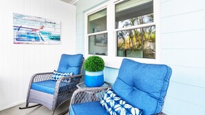 A porch with two wicker chairs, a blue cushion, a small potted plant, and a painting on the wall.