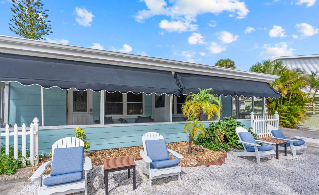 A beachfront property with a covered patio, white Adirondack chairs, and a small palm tree.