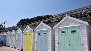 Une rangée de cabanes de plage colorées, chacune avec une porte numérotée, un escalier menant à une plateforme d’observation et un ciel dégagé.
