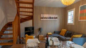 Un comedor con una escalera de madera, un sofá con cojines amarillos y una mesa preparada para seis personas, con copas de vino y platos.