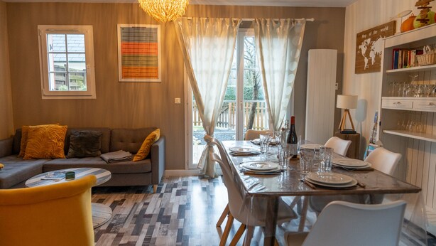 Un comedor con una mesa de madera dispuesta para cuatro, un sofá con cojines amarillos y una estantería con diversos objetos.