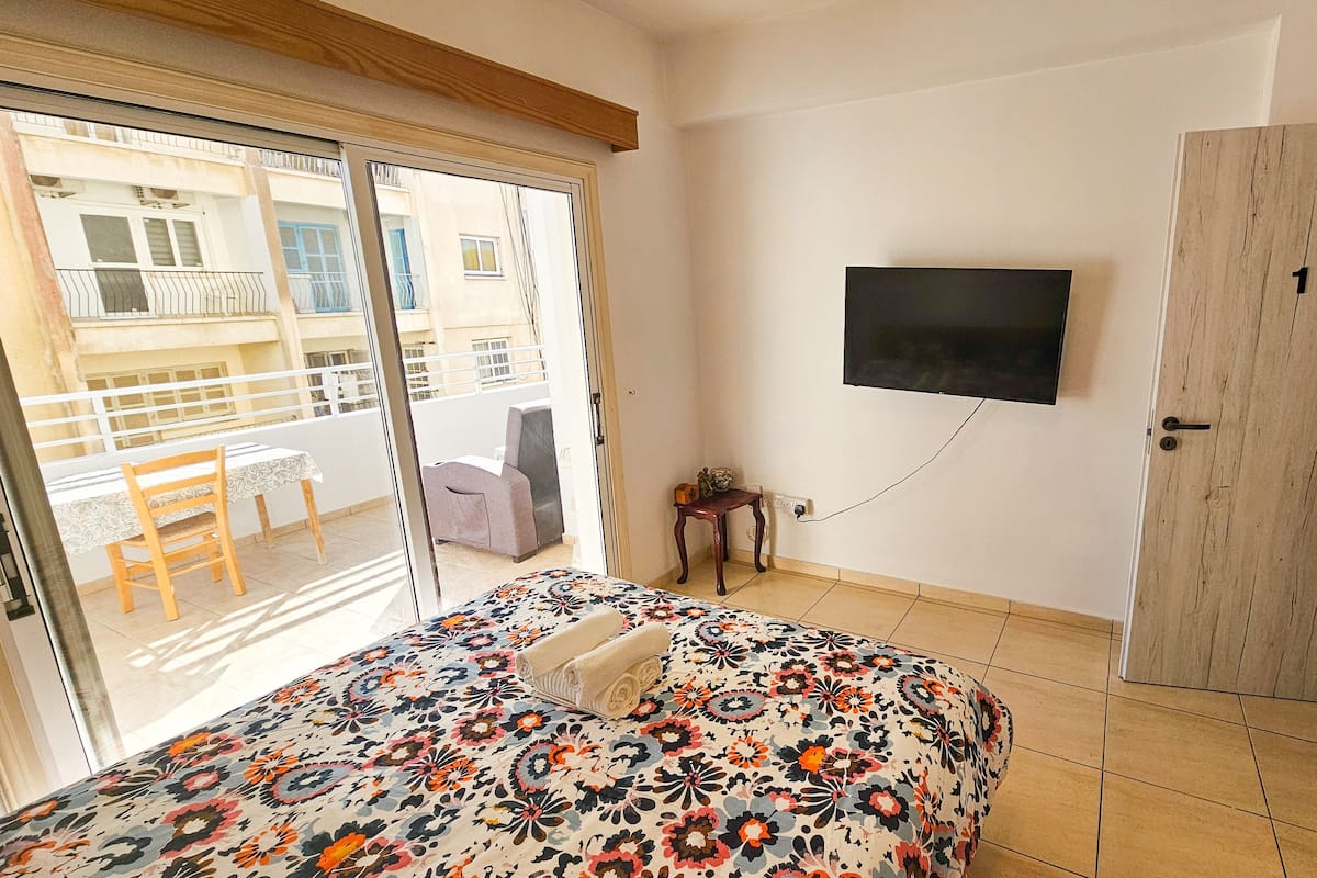 A bedroom with a bed, a TV mounted on the wall, a wooden door, and a balcony with a chair and table.
