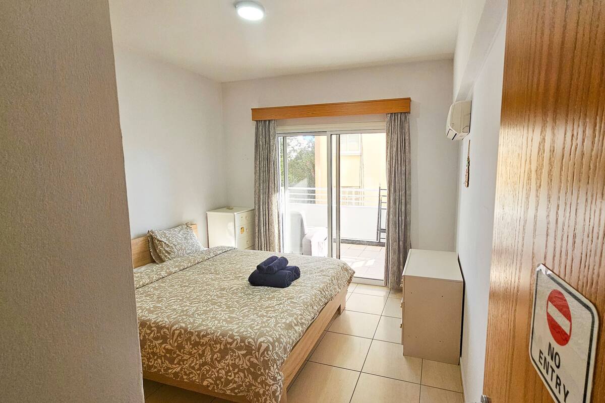 A hotel room with a bed, a nightstand, a suitcase, a balcony with a view, and a 'No Entry' sign.
