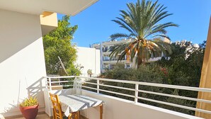 A balcony with a sofa, table, and chairs. There is a potted plant and a palm tree in the background. - Unique City Rooms (Larnaca)