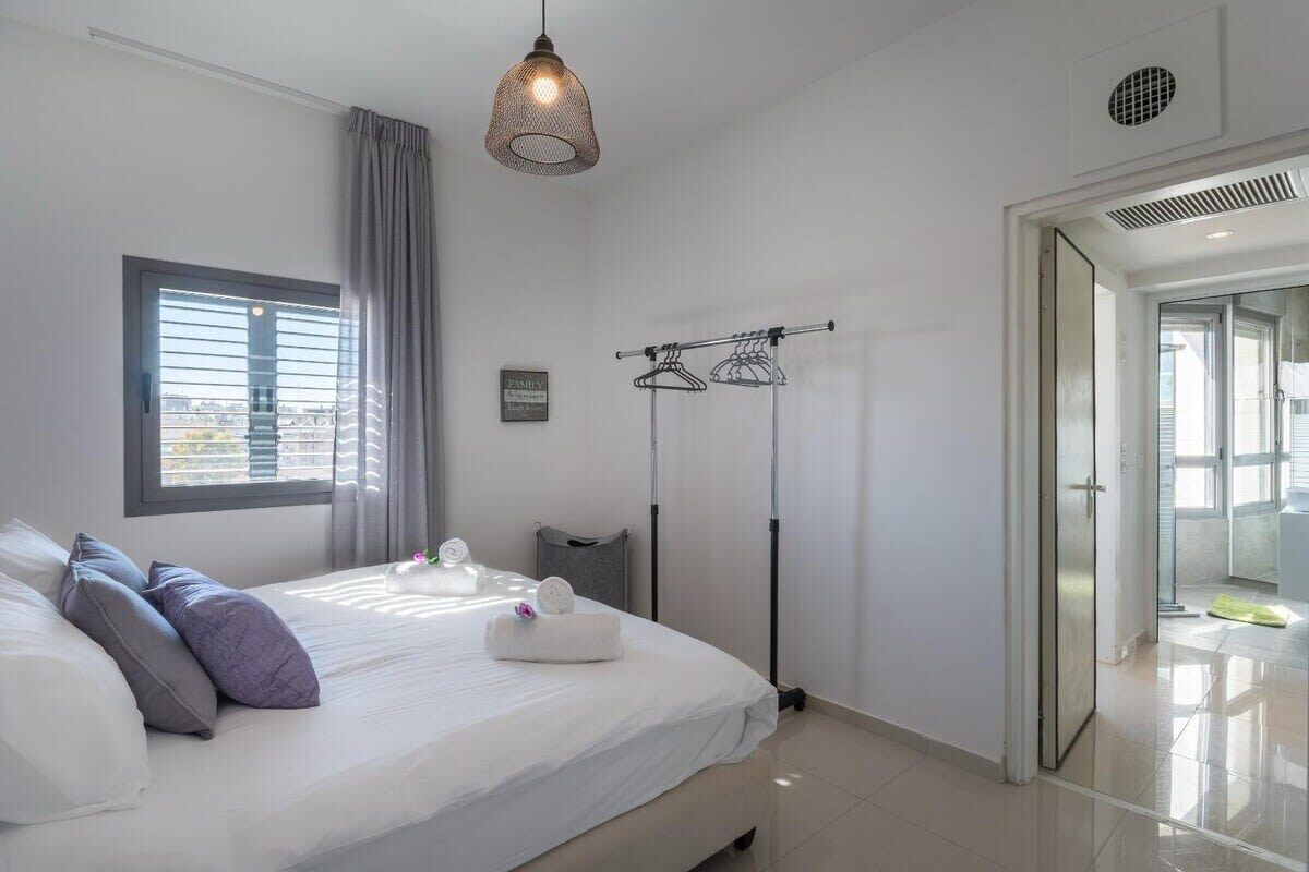 Une chambre moderne avec un grand lit, un luminaire suspendu, une fenêtre avec des stores et une porte donnant accès à une salle de bain.