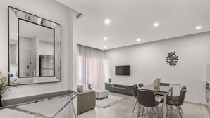 Una sala de estar moderna con comedor, un espejo grande, televisor de pantalla plana y una cocina.