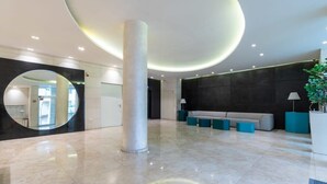Un vestíbulo amplio con suelo de mármol, una columna central imponente, un espejo circular, un sofá y una planta en maceta.