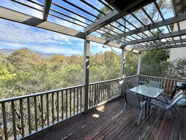 A wooden deck with a table and chairs, overlooking a forested area with mountains in the distance.