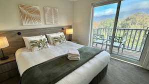 A hotel room with a bed, two wall art pieces, a nightstand with a lamp, and a balcony with outdoor furniture.