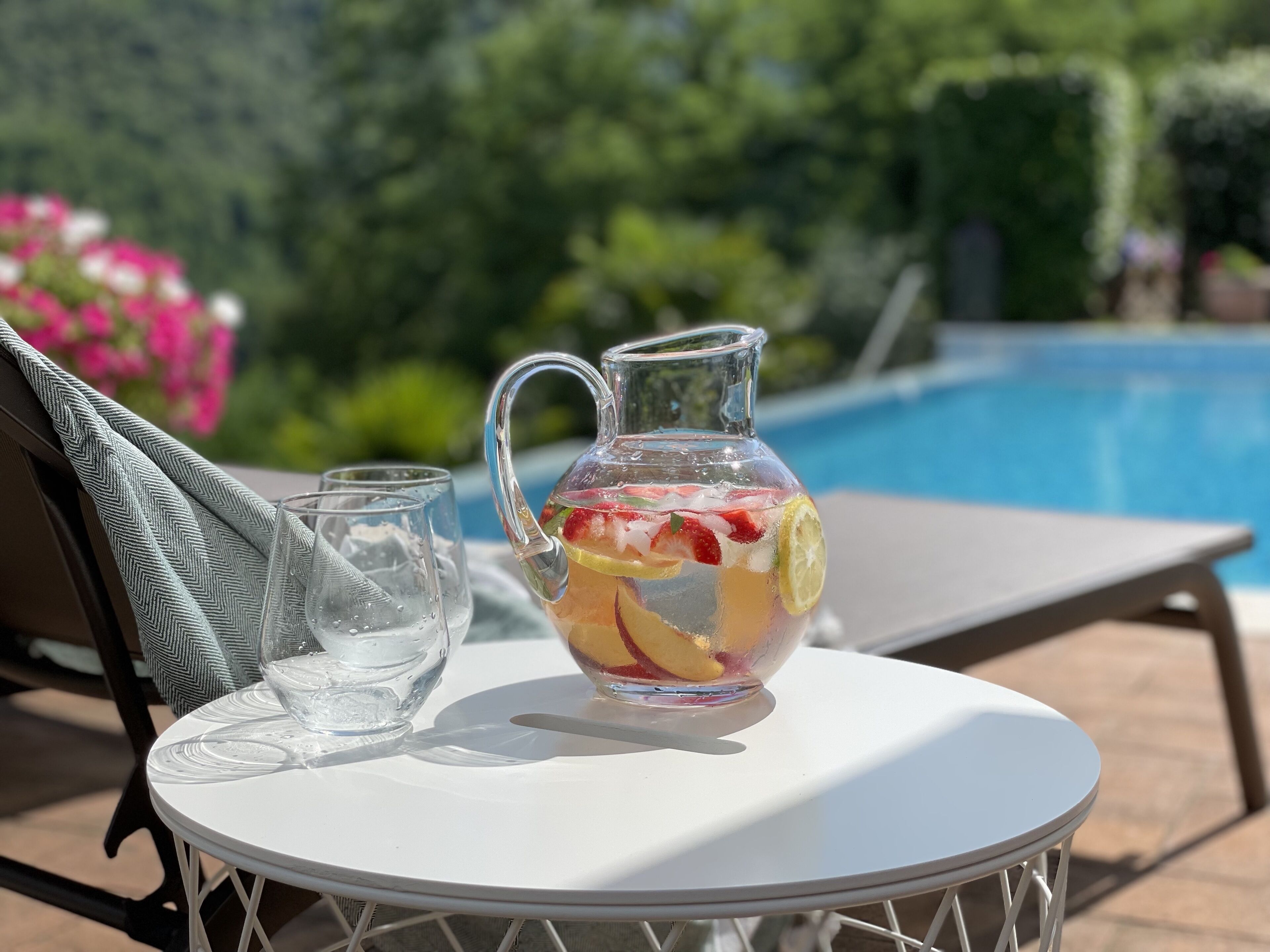 A clear glass pitcher with fruit and ice on a white table by a pool.