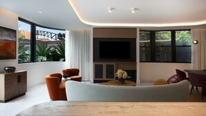 A modern living room with a large TV, comfortable seating, and large windows offering a view of greenery. - Kirribilli Charisma I90 (Kirribilli)