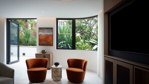 A modern living room with two orange chairs, a small side table, and a painting on the wall. Large windows provide views of greenery outside. - Kirribilli Charisma I90 (Kirribilli)