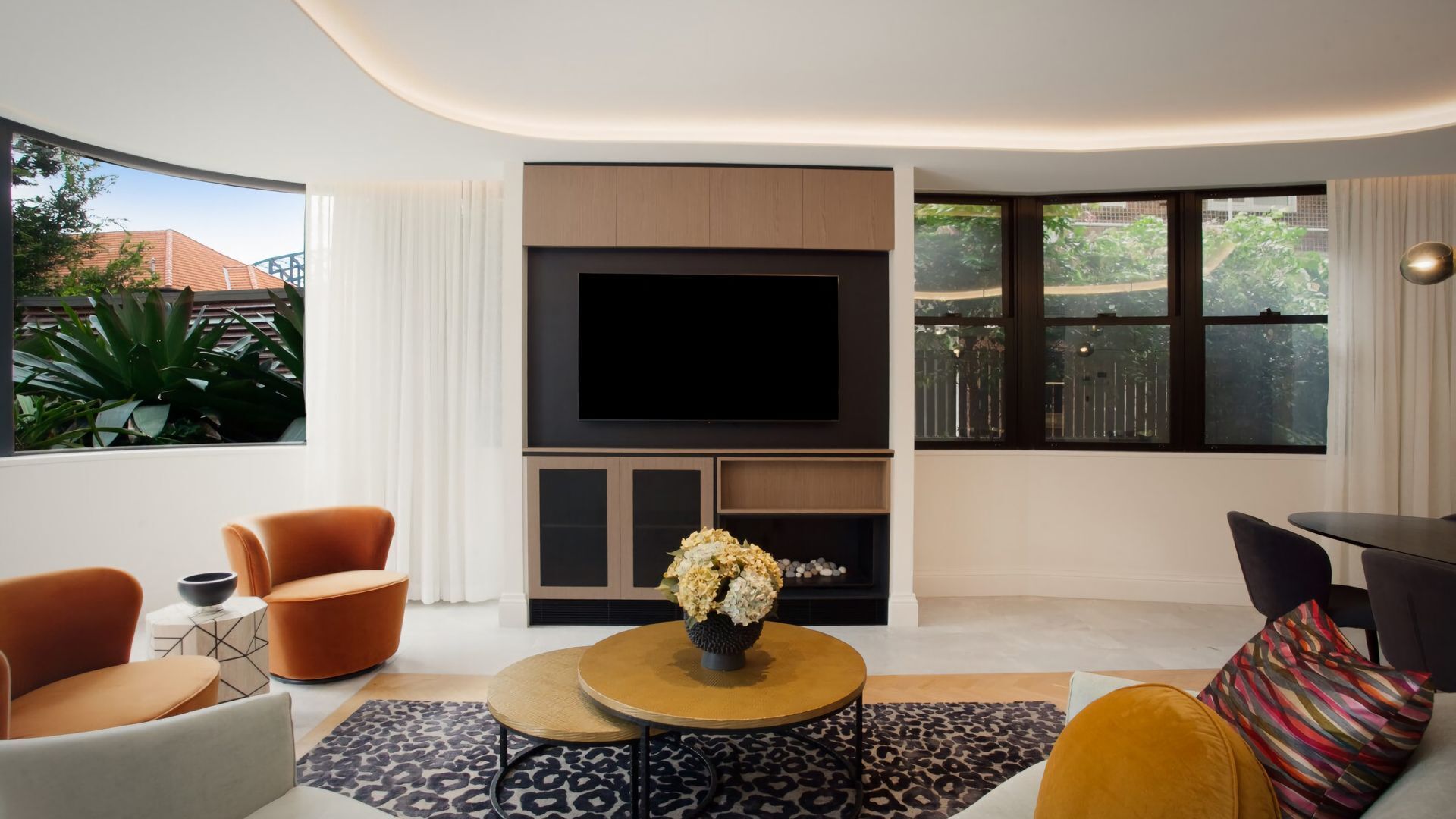 Ein modernes Wohnzimmer mit Flachbildfernseher, bequemen Sitzmöglichkeiten und großen Fenstern mit Blick auf Grünflächen.