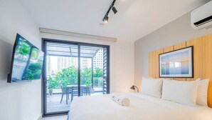 A modern hotel room with a large bed, a TV mounted on the wall, and a sliding glass door leading to an outdoor area with a table and chairs.