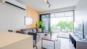 A modern living room with a sofa, coffee table, and TV. There is a large window with a view of buildings and greenery outside.