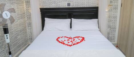 A neatly made bed with white linens and a heart-shaped decoration of rose petals.