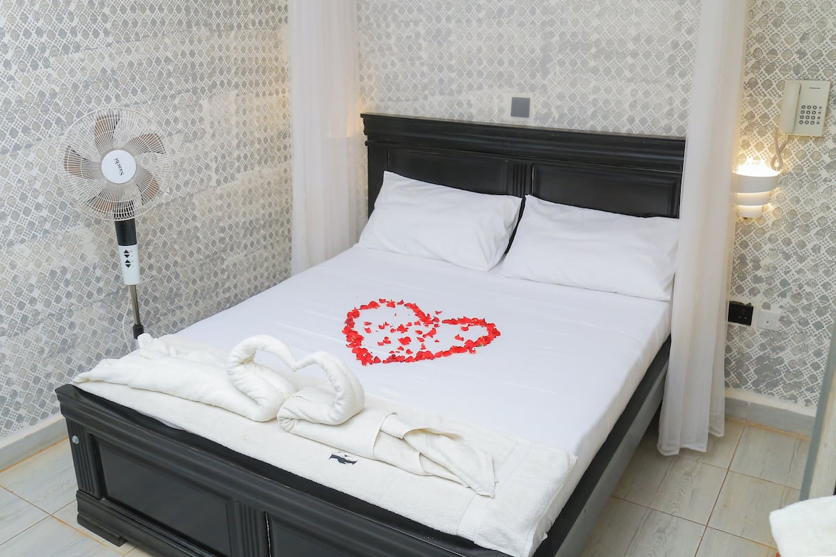 A neatly made bed with white linens, a heart-shaped decoration on the bedspread, and a fan beside the bed.