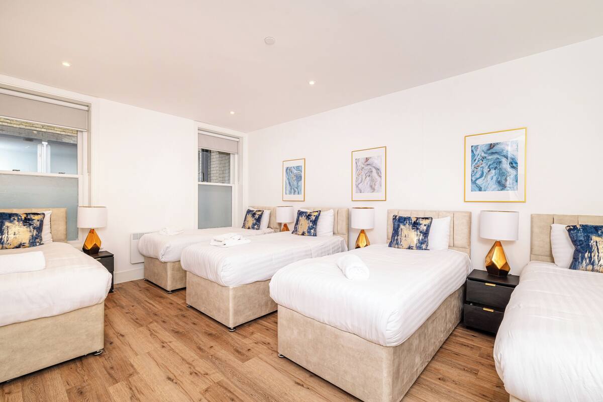 Habitación de hotel con cuatro camas, cada una con edredón blanco y almohadas a juego. Se observan mesitas de noche con lámparas y un estante en la pared.