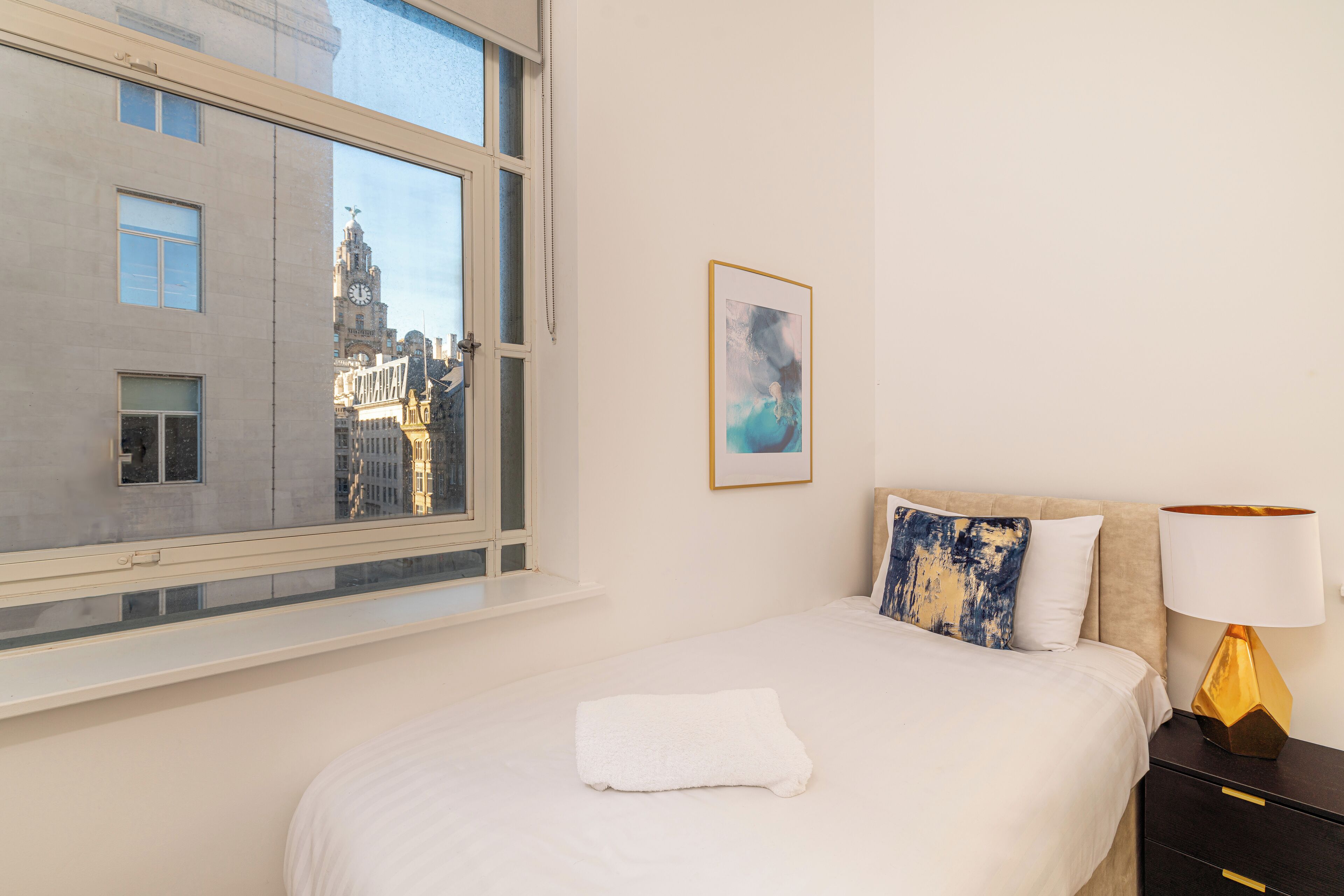 Une chambre d’hôtel avec une grande fenêtre, un lit avec une literie blanche, un tableau au mur et une table de chevet avec une lampe.