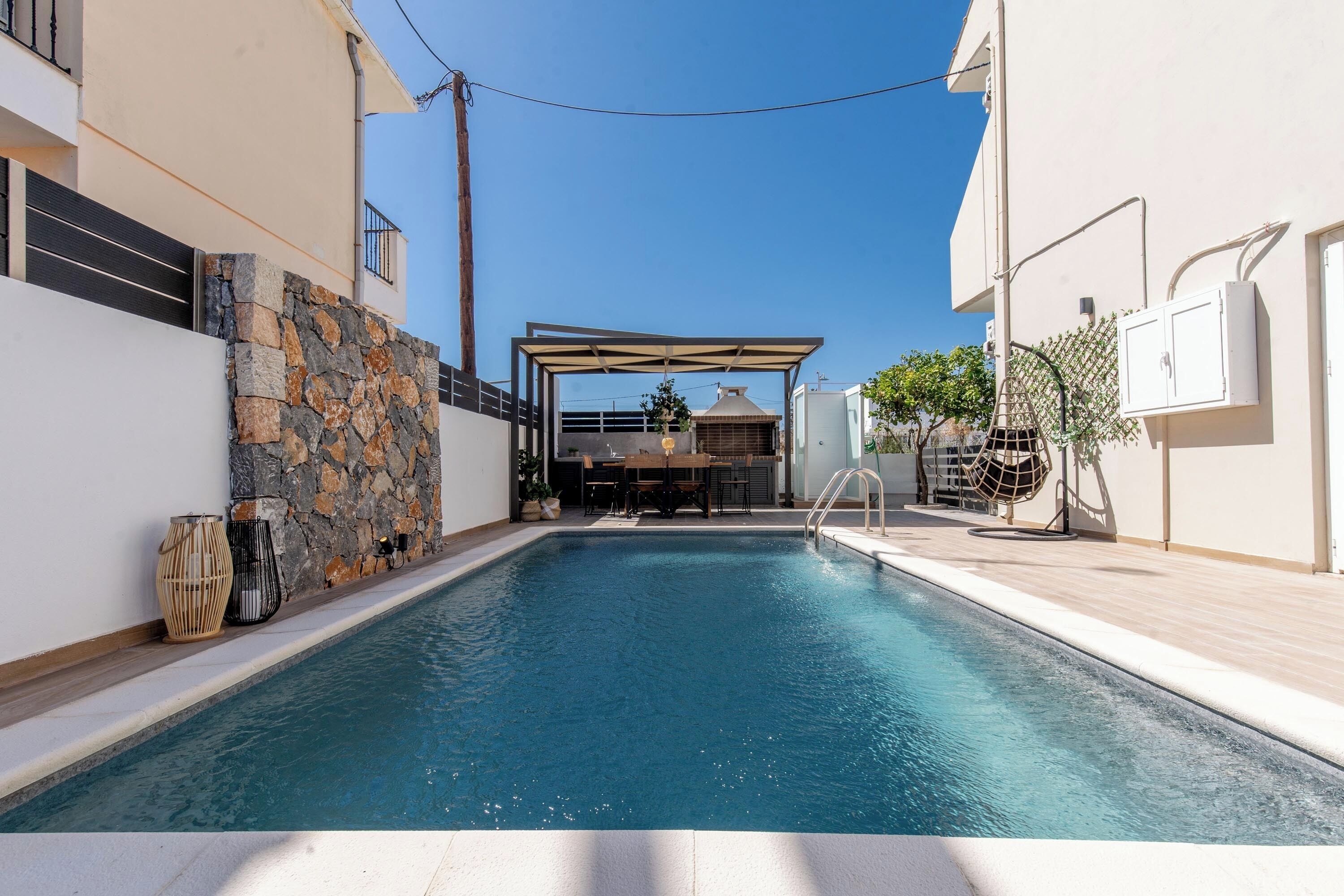 A swimming pool with a stone wall on one side, a covered outdoor area, and a building with a balcony.