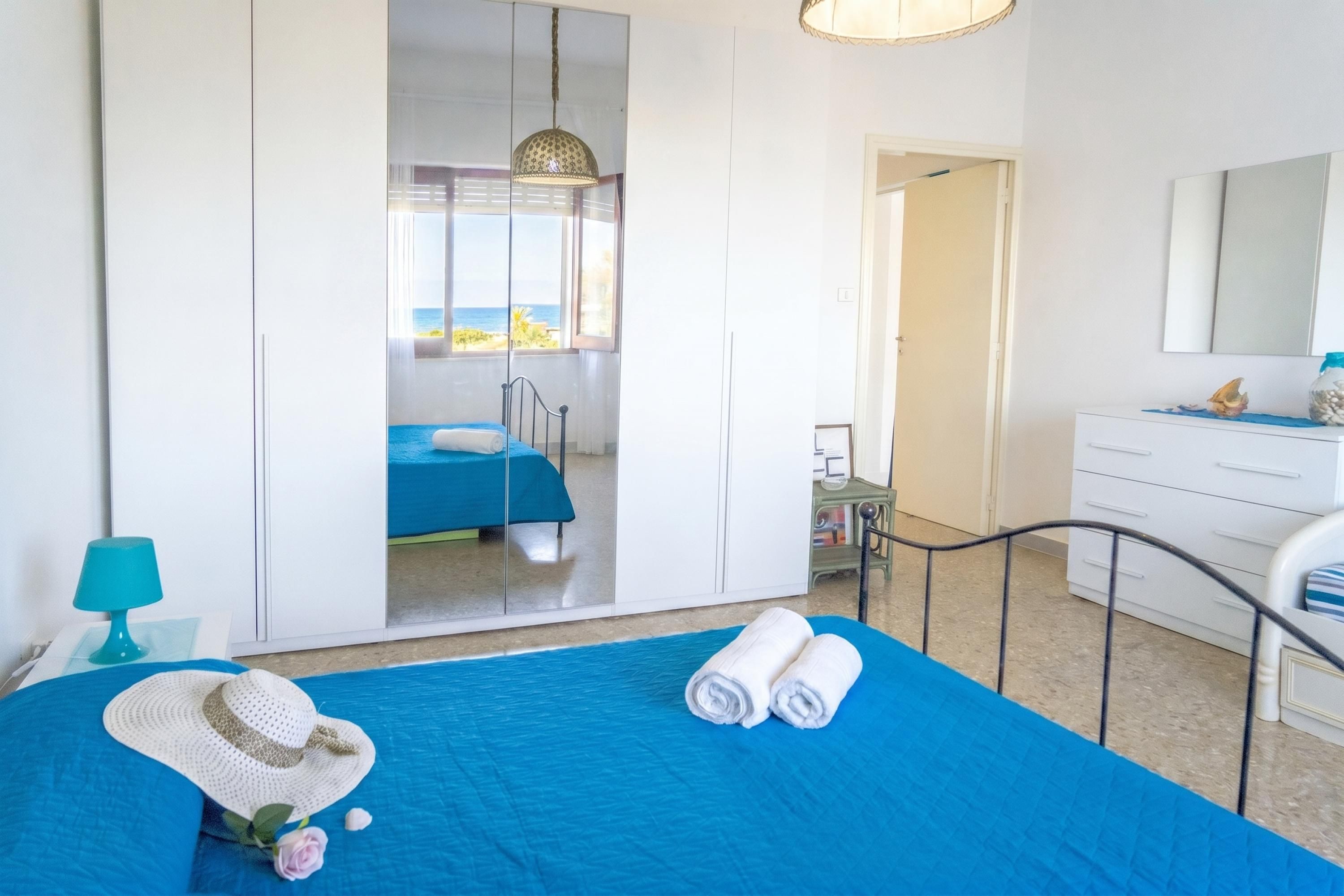 A bedroom with a bed, a mirror, a dresser, and a view of the sea.