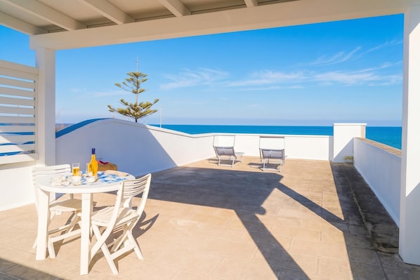 Une terrasse avec une table et des chaises, une bouteille et un verre, donnant sur une mer d’un bleu limpide.