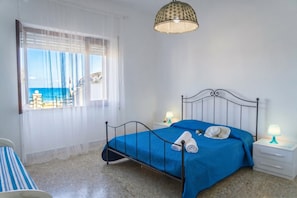 Quarto com cama, mesa de cabeceira, luminária, janela com cortinas e vista para o mar.