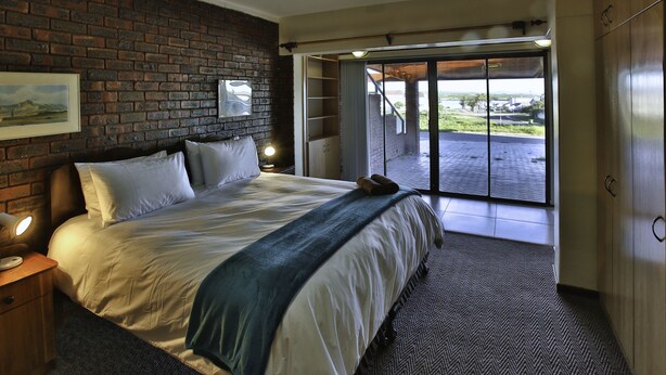 A bedroom with a large bed, a brick wall, a nightstand, a lamp, and a view through a sliding door to an outdoor area. - Dasklip Langebaan 8-sleeper (Langebaan)