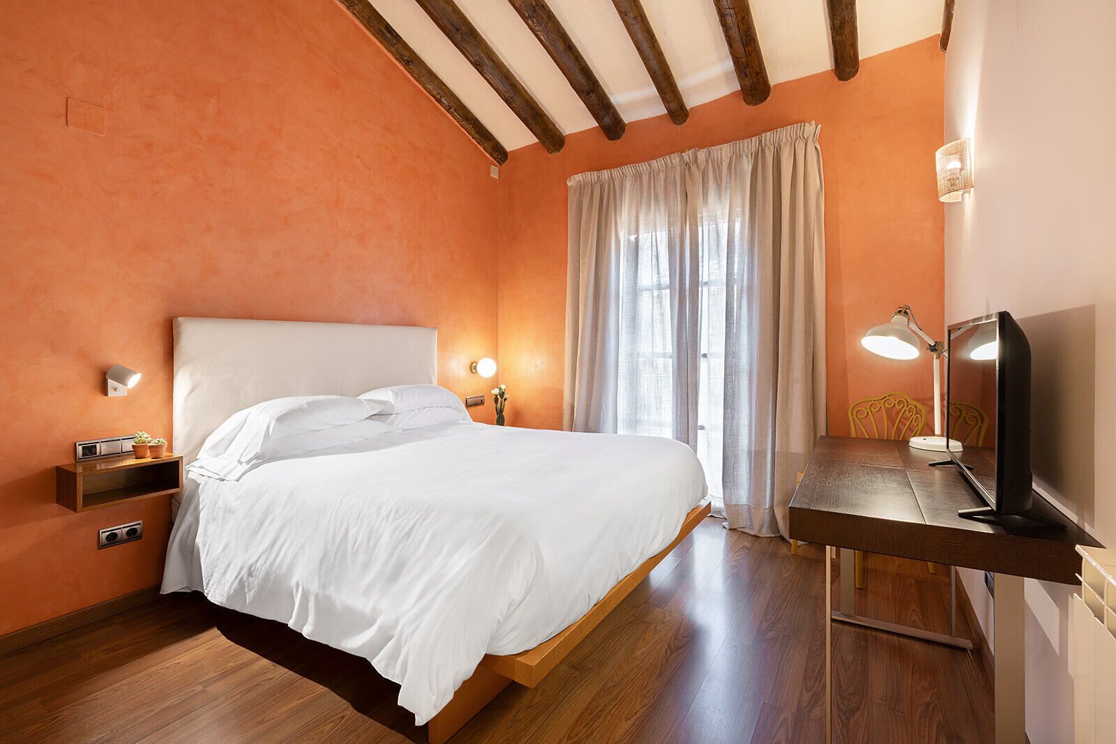 A bedroom with a wooden bed, a nightstand with a lamp, a desk with a computer, and a window with curtains.