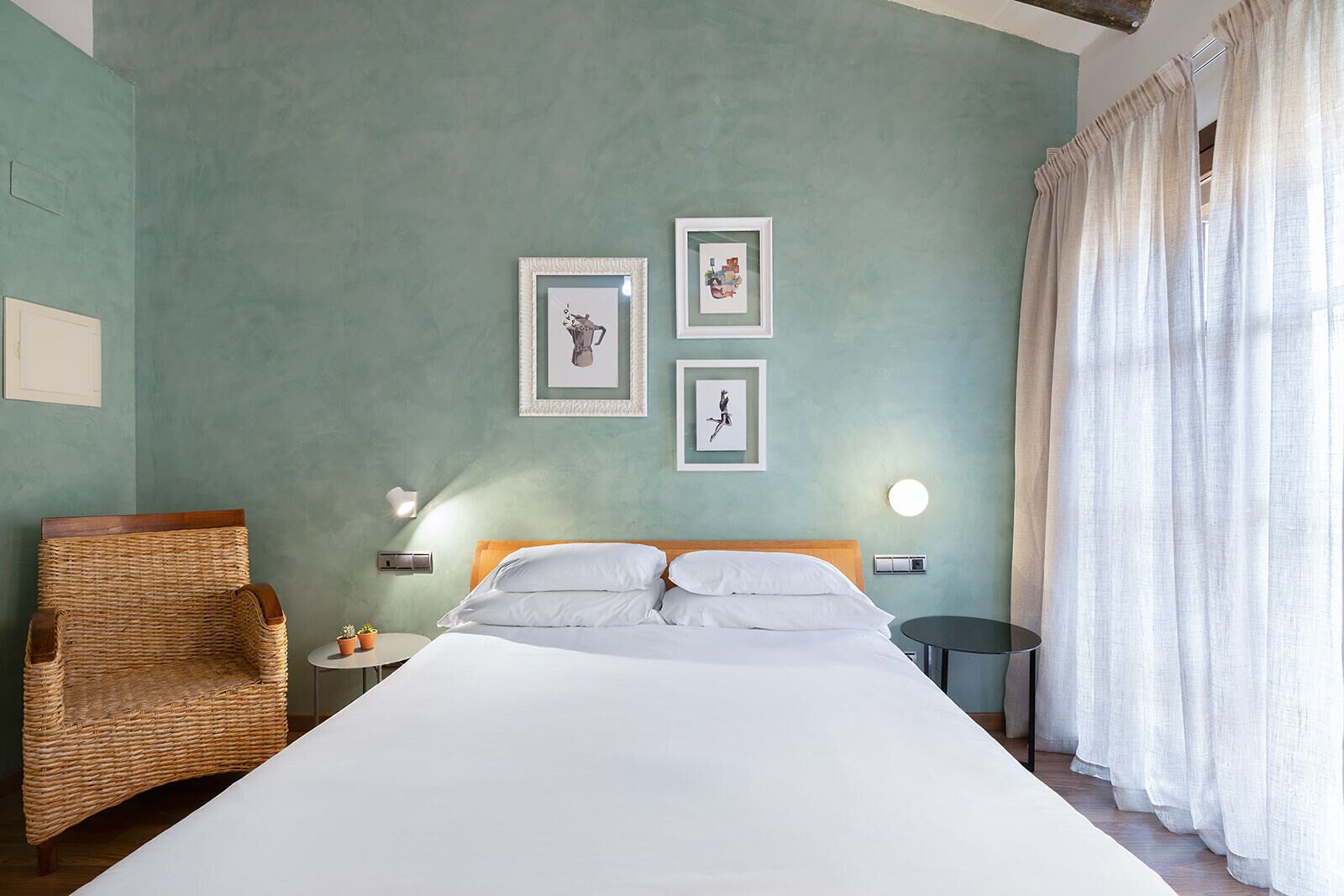 A bedroom with a large bed, a wicker chair, two framed pictures on the wall, a round table, and a window with curtains.