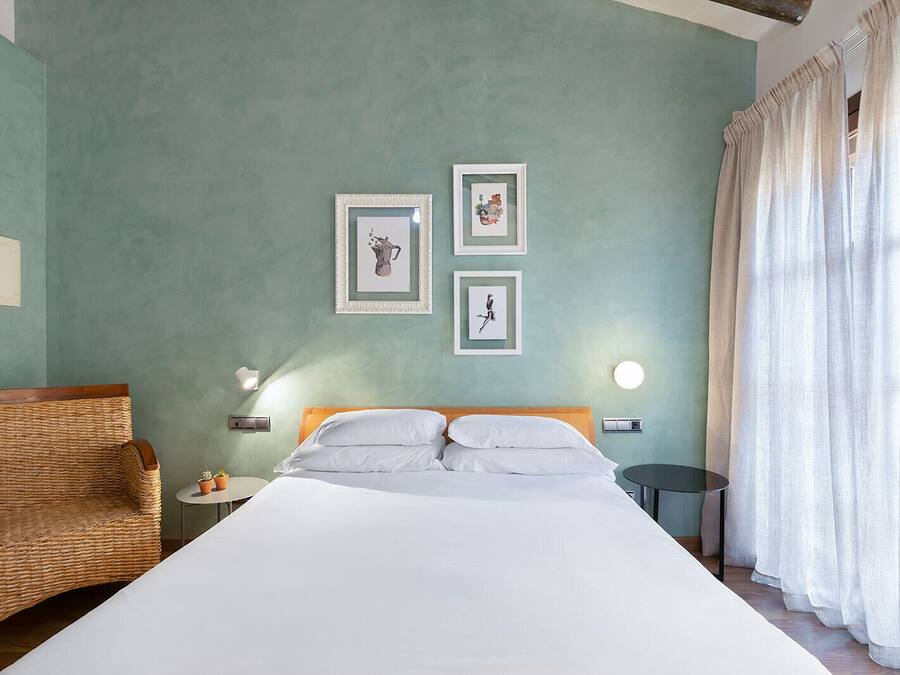 A bedroom with a large bed, a wicker chair, two framed pictures on the wall, a round table, and a window with curtains.
