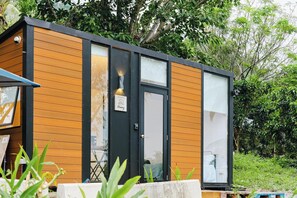 Une cabane moderne en bois, à deux étages, dotée de grandes fenêtres et d’une porte noire, et entourée d’une végétation luxuriante.