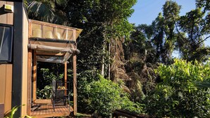 A wooden structure with a covered entrance, a bench, and a table, surrounded by dense greenery and a rocky cliff. - Magic Mena Rainforest Retreat by Tiny Away (Mena Creek)