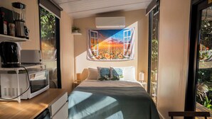 A bedroom with a bed, a desk with a microwave, a coffee maker, and a window with a view of a field. - Magic Mena Rainforest Retreat by Tiny Away (Mena Creek)