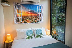 Une chambre avec une grande fenêtre, un lit recouvert d’une literie blanche, deux coussins décoratifs, une table de chevet avec une lampe et une tapisserie murale.