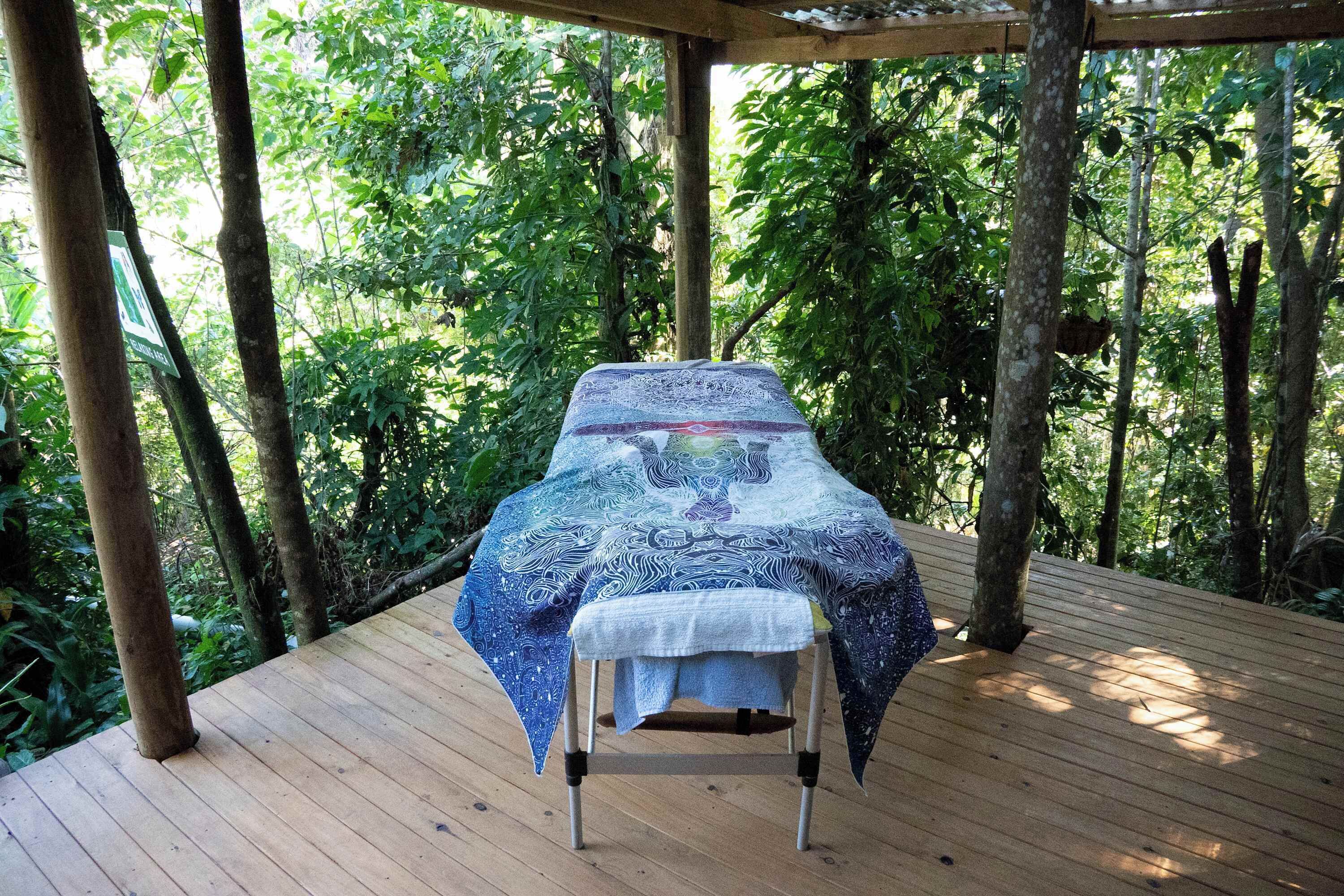Een houten terras met een tafel, afgedekt met een patroonkleed, omringd door weelderige begroeiing.