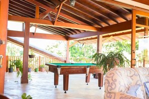 A pool table in a covered outdoor area with wooden beams, potted plants, and a patterned sofa.