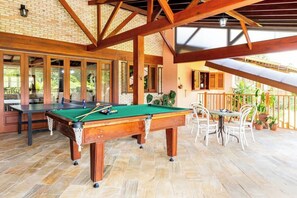 A pool table with a green felt surface, wooden legs, and a white cloth. There is a seating area with white chairs and a table to the right. The room has a brick wall and a wooden ceiling structure.