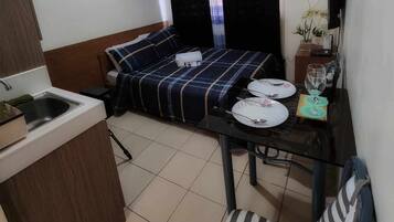 A compact hotel room with a bed, a small table with plates and glasses, a sink, and a window with curtains.