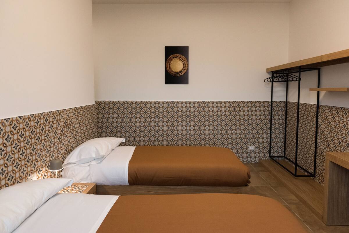 Une chambre d’hôtel avec deux lits, un papier peint à motifs, une œuvre d’art accrochée au mur, une table de chevet avec une lampe et un bureau en bois avec un porte-manteau en métal.