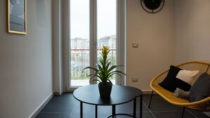 Een moderne woonkamer met een ronde tafel, een potplant, een rieten stoel met kussens en een wandklok.