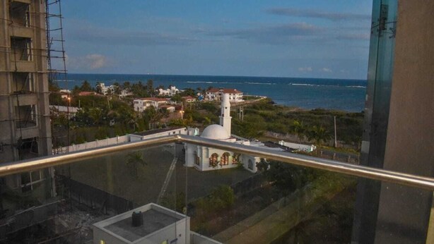 Blick aus einem Fenster auf eine Küstenlandschaft mit einem weißen Gebäude, das eine Kuppel und einen Turm hat, umgeben von Grünflächen und Wohnhäusern.