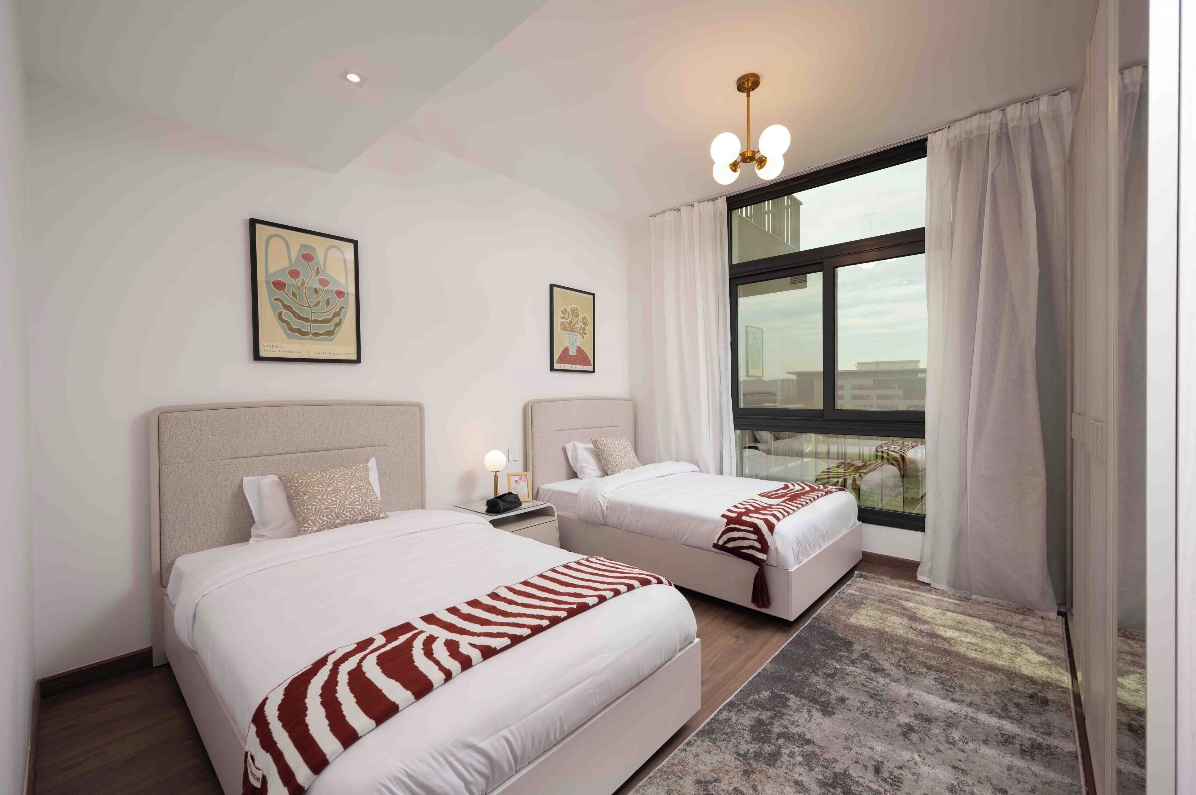A hotel room with two beds, a large window with curtains, and artwork on the walls.