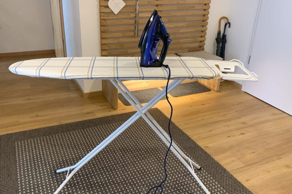 Una plancha compacta con un hierro azul, un banco de madera y una pared blanca al fondo.