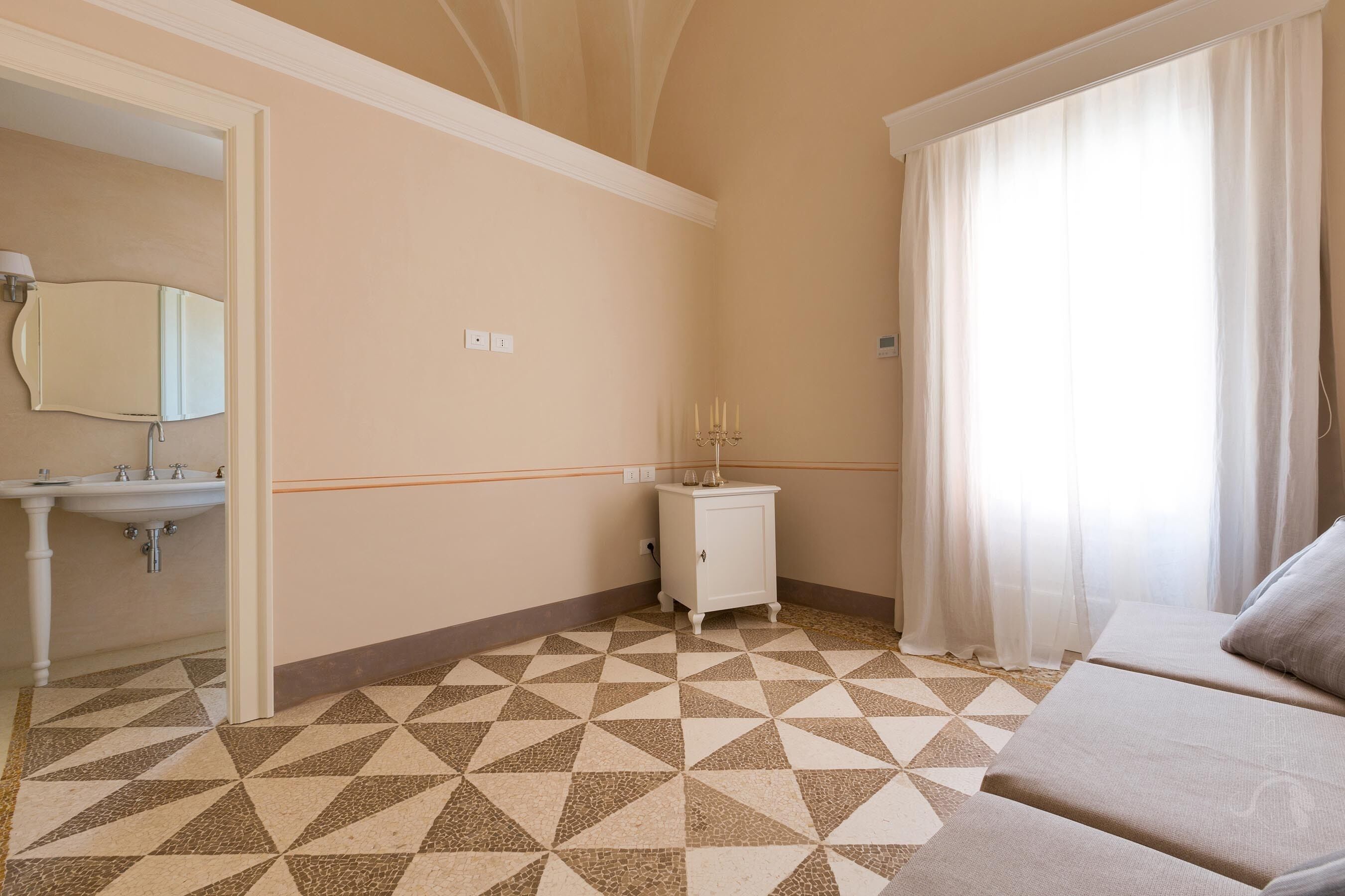 A hotel room with a checkered floor, a bed, a small bedside table, a mirror, and a window with curtains.
